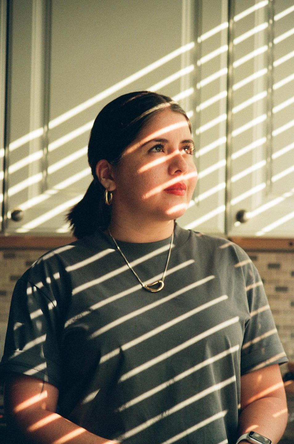 A woman with a necklace and earrings looks up, with shadows from blinds cast across her face and body.