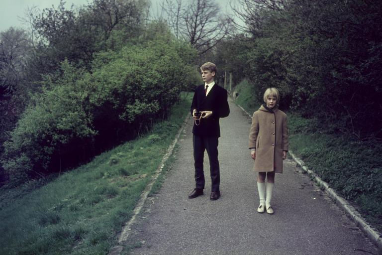 A boy and a girl stand on a paved path surrounded by green foliage.