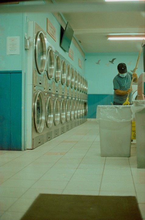 A man wearing a face mask sweeps the floor of a laundromat with a row of washing machines.