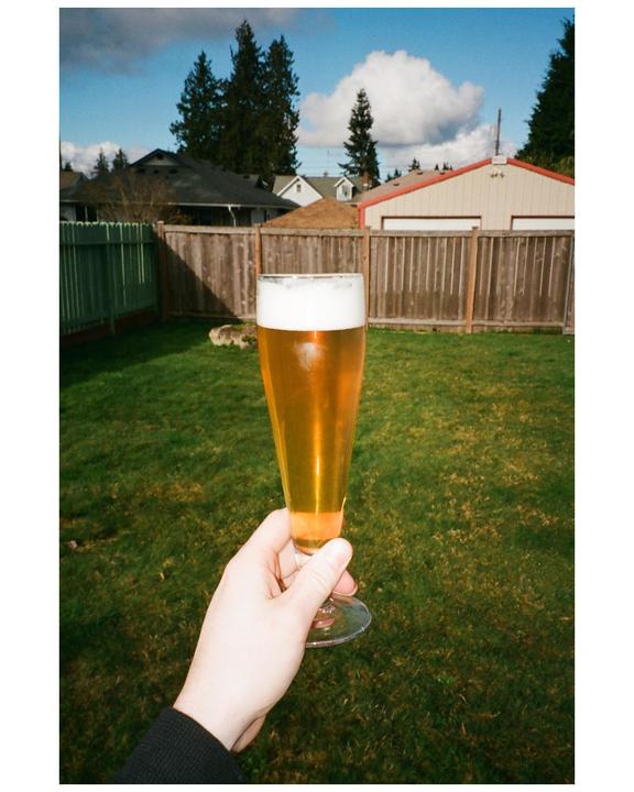 A hand holds a glass of beer in a backyard with houses and trees in the background.