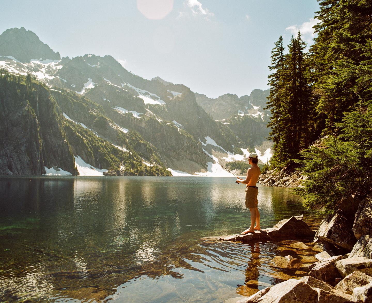A man stands barefoot on a rock at the edge of a clear mountain lake, fishing with a rod, with snow-capped mountains and pine trees in the background.