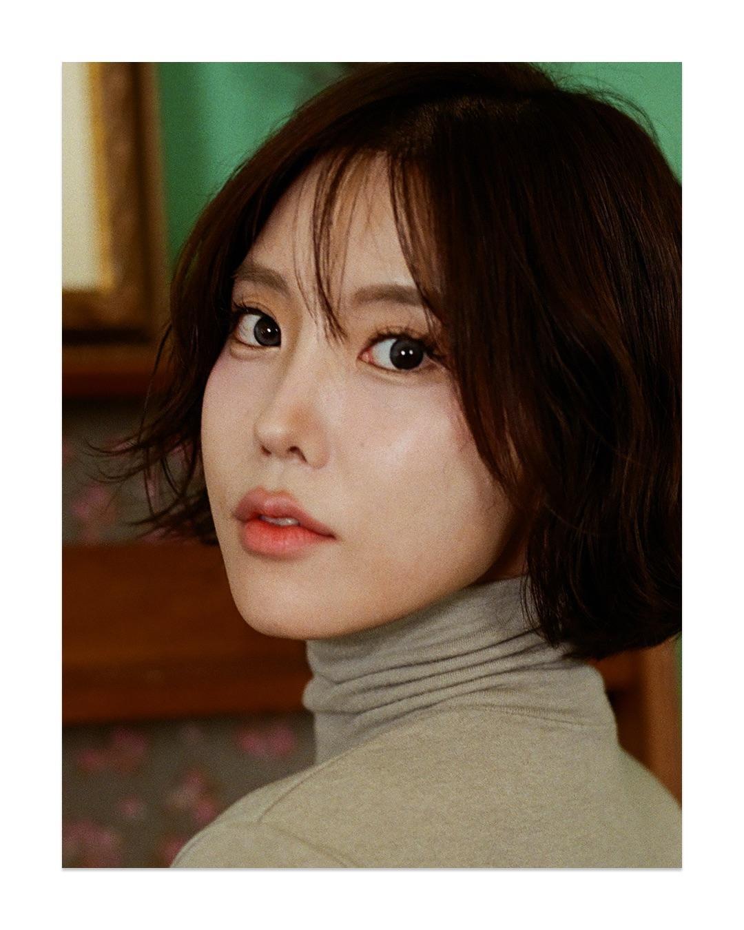 A woman with short hair and a turtleneck sweater looks over her shoulder at the viewer.