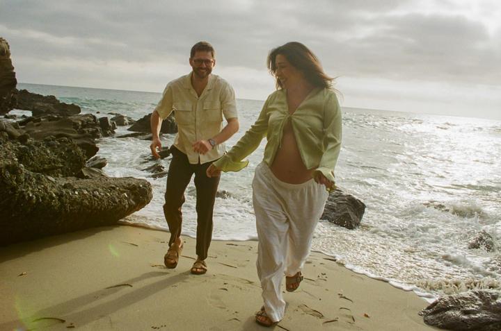 A couple runs along a sandy beach with rocks and ocean waves.
