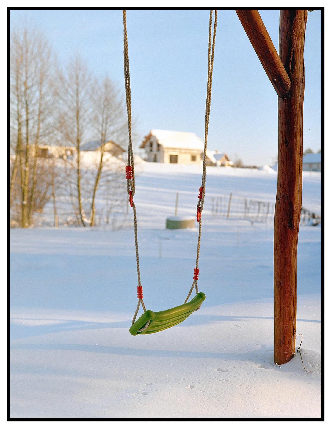 A green swing hangs from a wooden frame in a snowy landscape with houses and trees in the background.