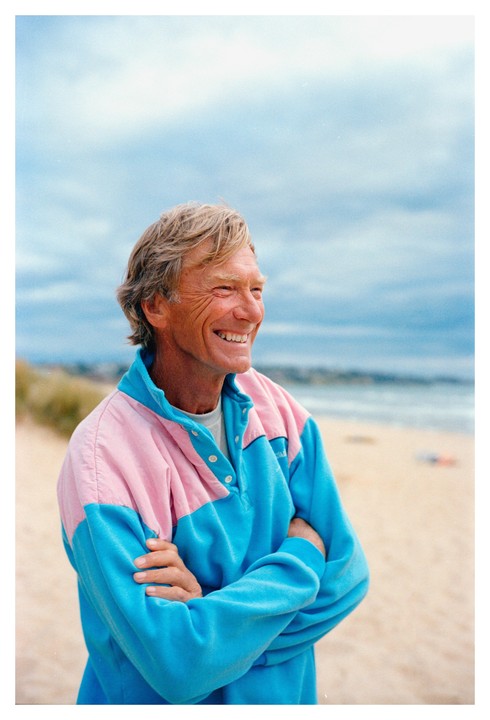 A man with blond hair and a pink and blue sweatshirt smiles while standing on a beach.