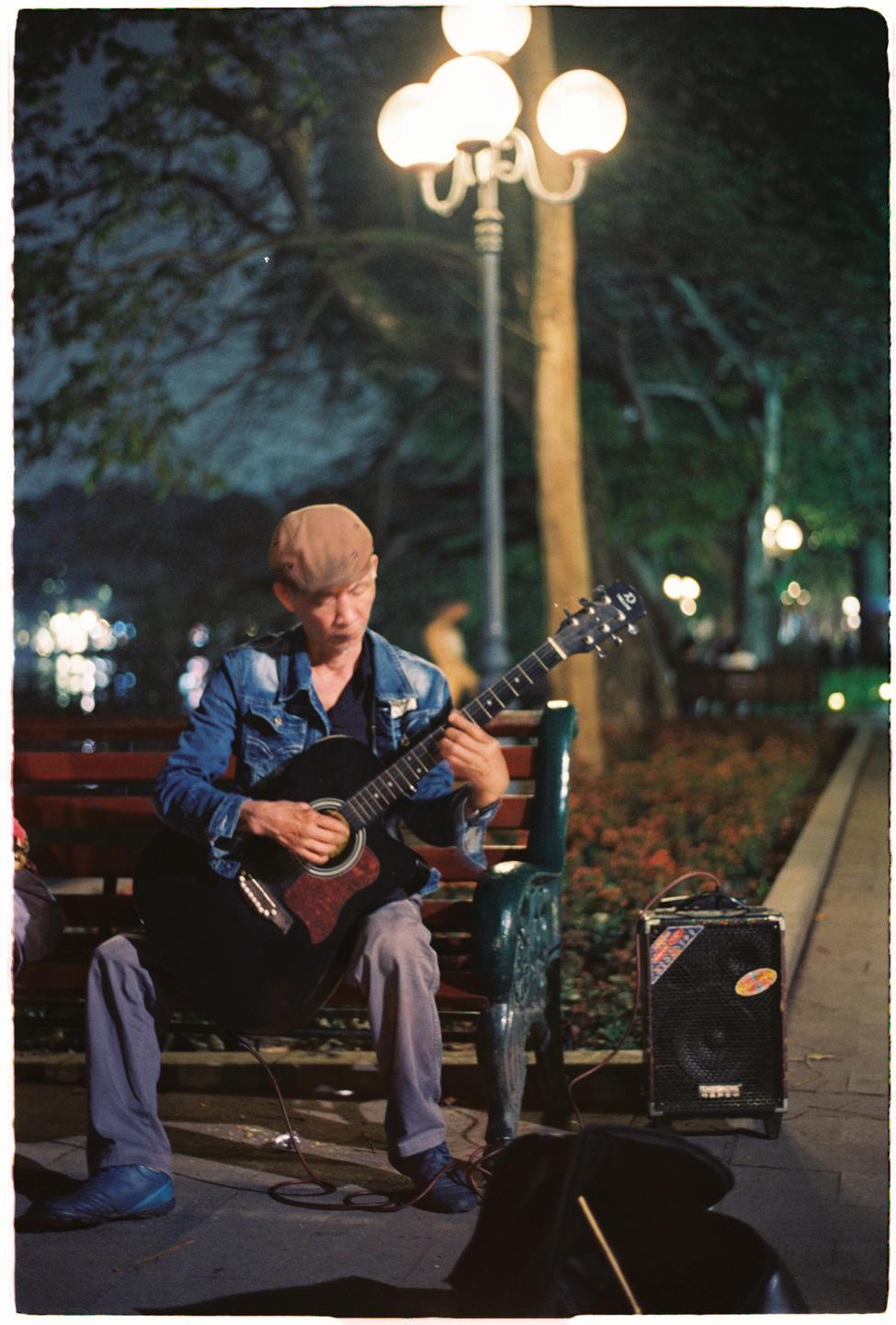 A man wearing a denim jacket and a cap sits on a park bench playing an acoustic guitar at night.
