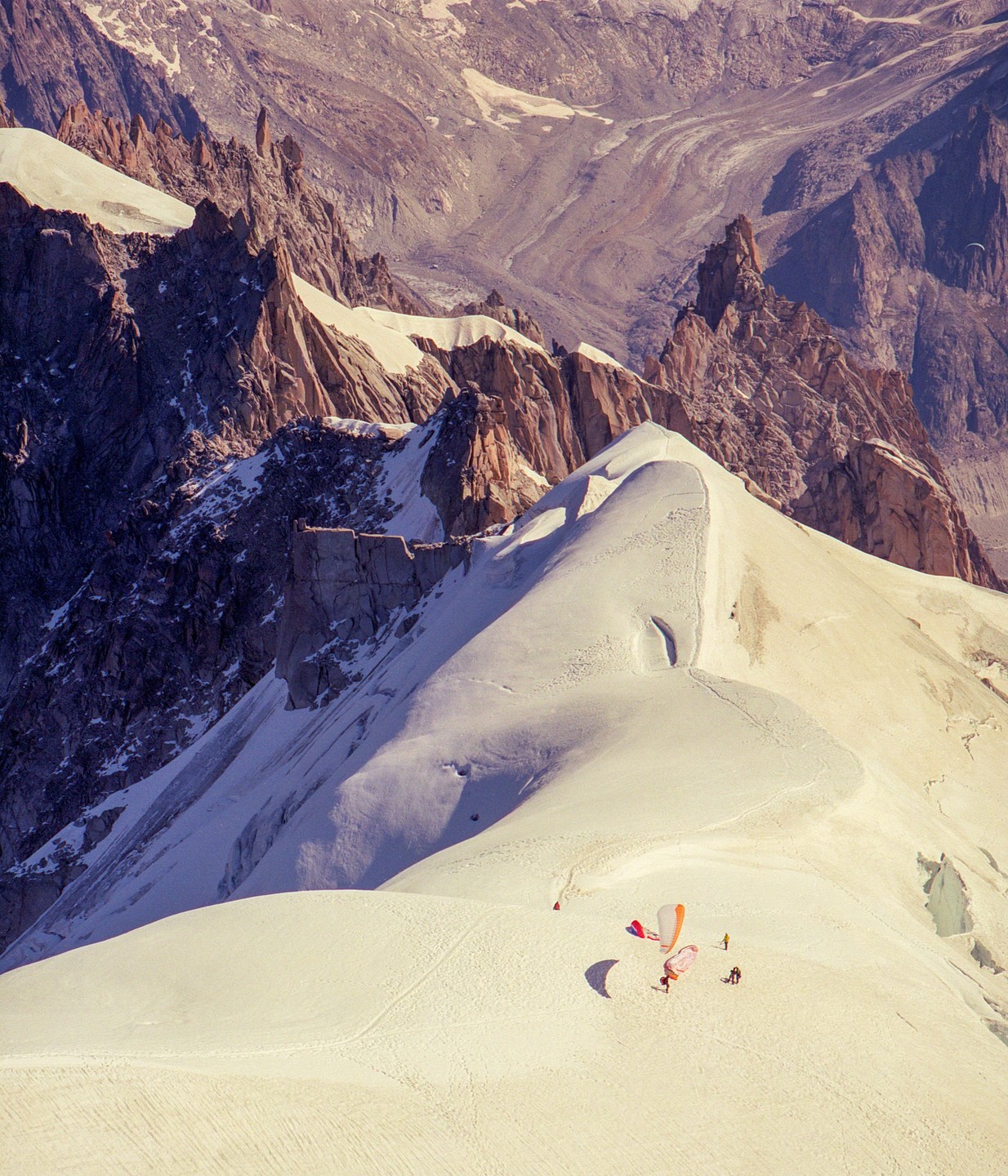 Paragliders are preparing to launch from a snowy mountain ridge with a vast mountain range in the background.