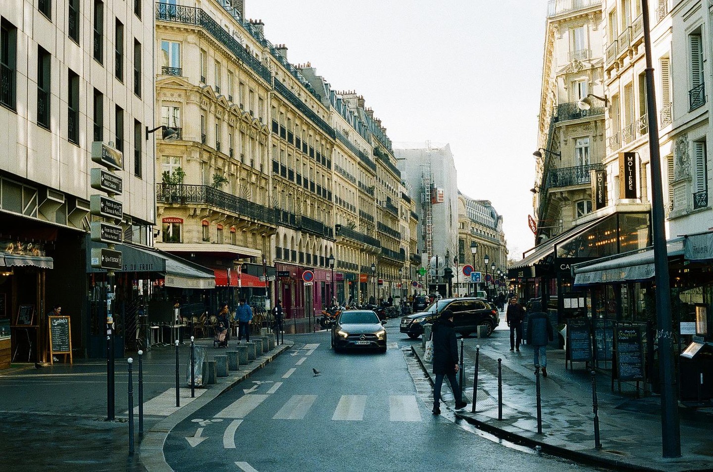 A street in Paris is lined with buildings and shops, with cars and people moving along the wet pavement.