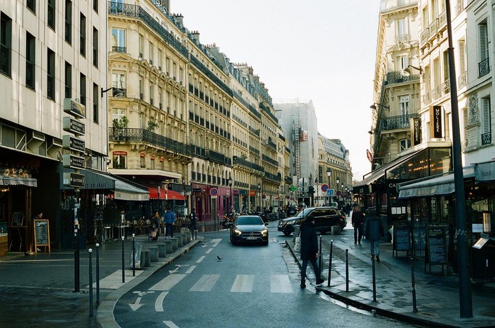 A street in Paris is lined with buildings and shops, with cars and people moving along the wet pavement.