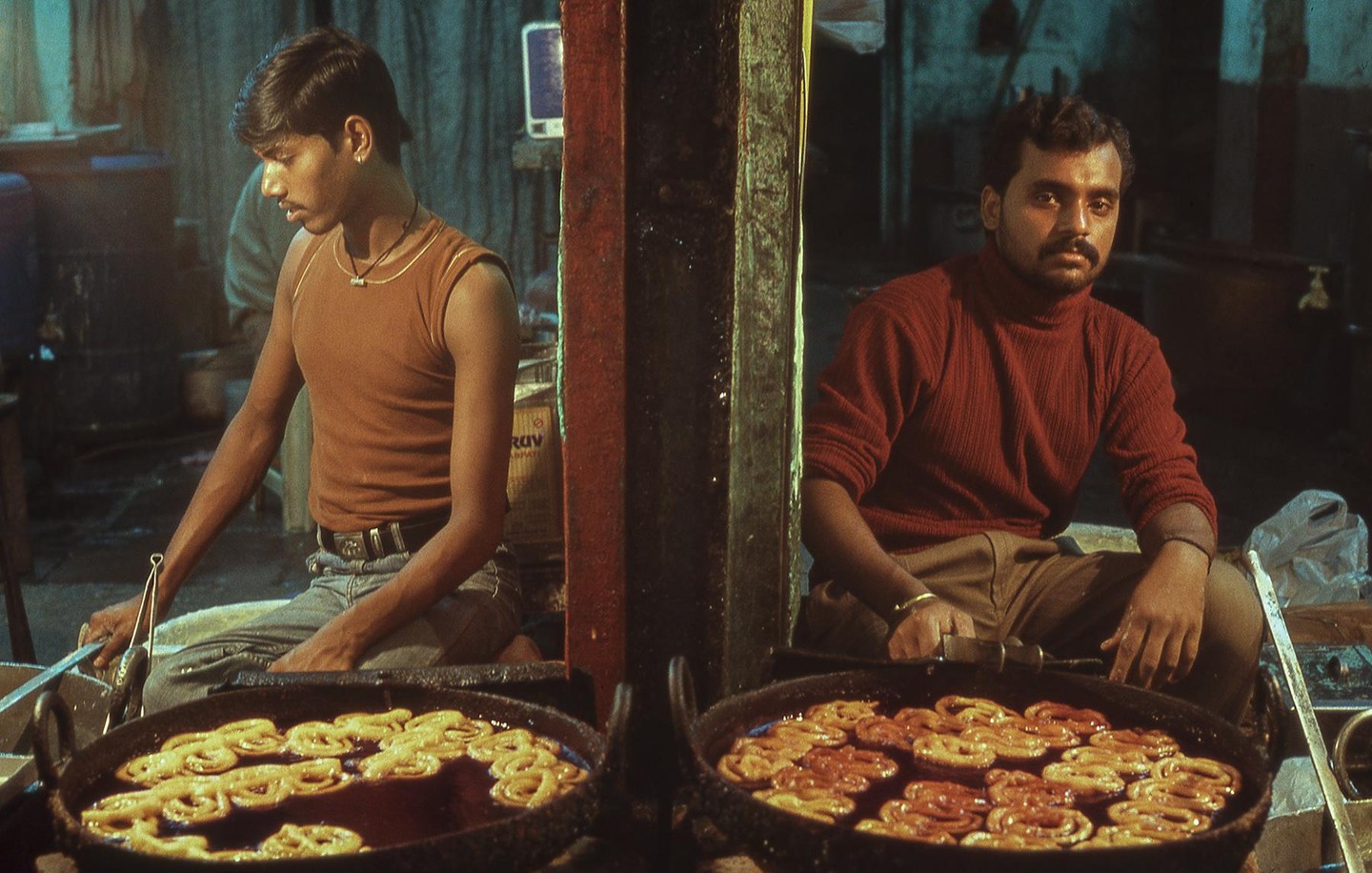 Two men sit on the ground next to large pans of jalebi frying in oil.