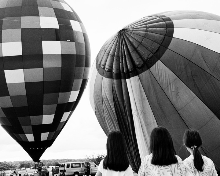 Three women stand with their backs to the camera, looking at two hot air balloons on the ground.