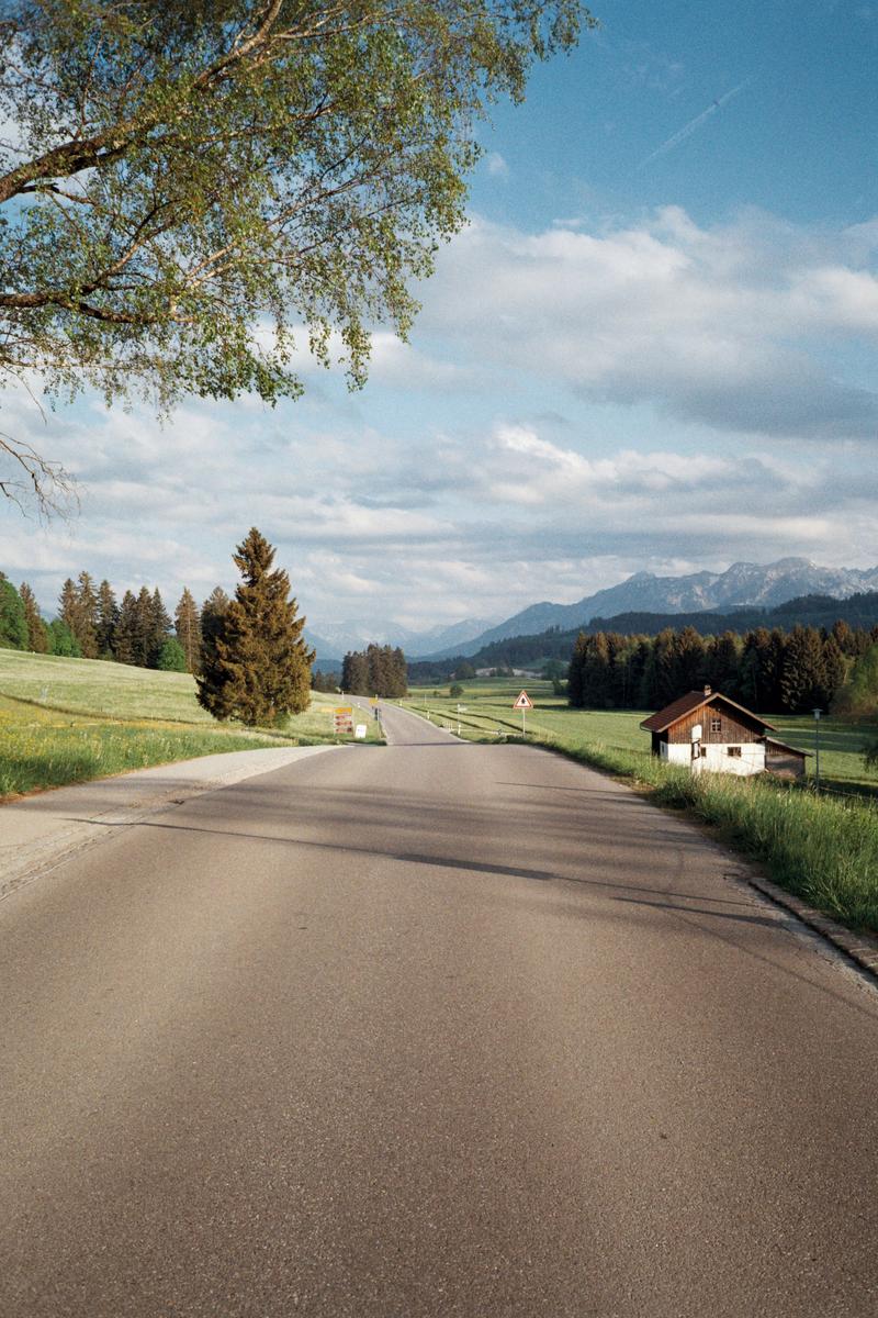 A long road stretches into the distance, flanked by green fields, trees, and mountains under a cloudy sky.