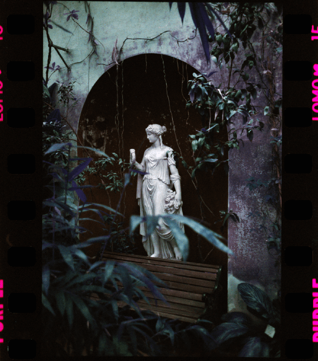A statue of a woman stands in a garden archway surrounded by plants.