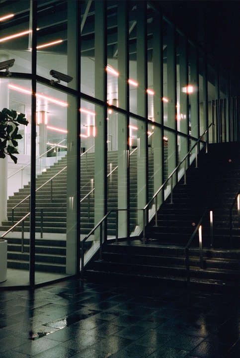 A modern building with glass walls and multiple staircases is illuminated by neon lights at night.