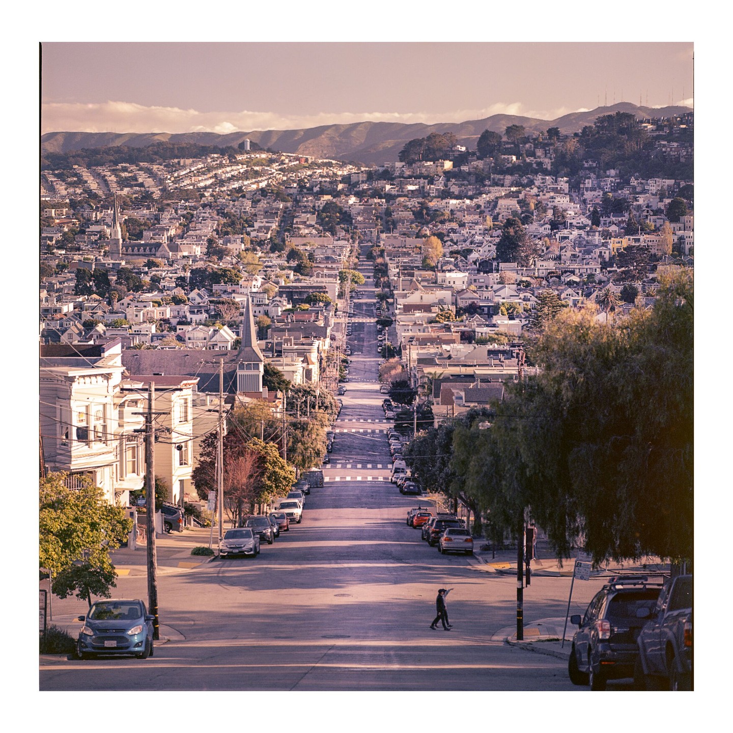 A person walks across a street that stretches down a hill through a city with houses and mountains in the background.