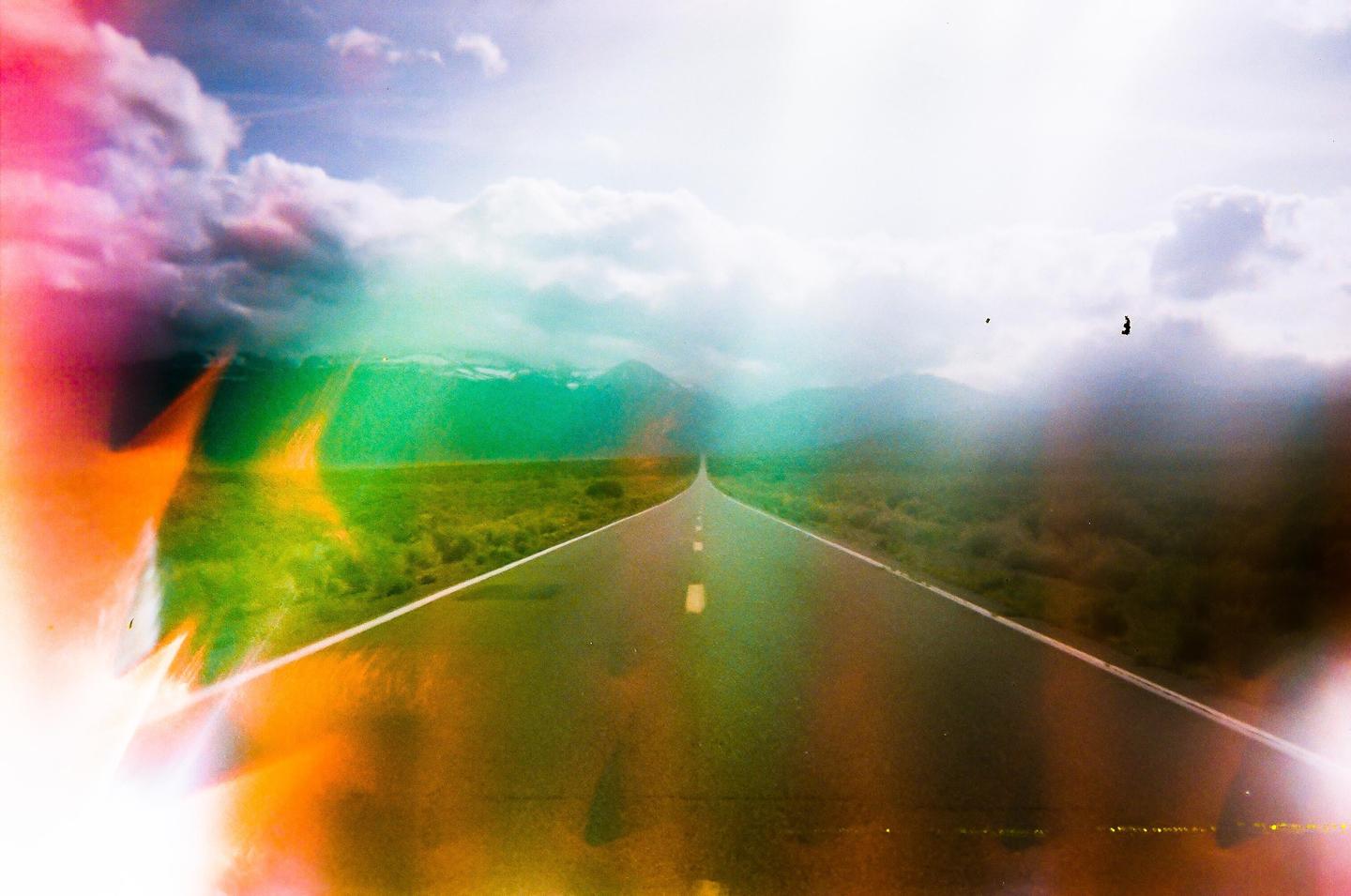 A long, straight road extends into the distance, flanked by green fields and mountains under a cloudy sky, with colorful light leaks on the left and bottom.