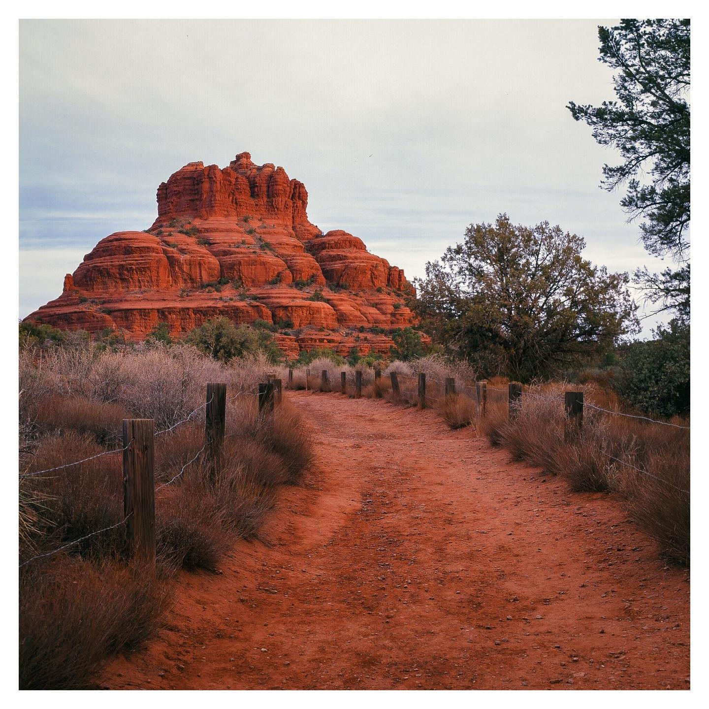A red dirt path with a barbed wire fence leads towards a large red rock formation under a cloudy sky.