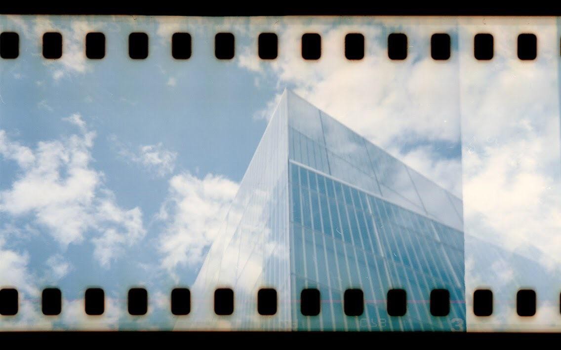 A glass building reflects a cloudy blue sky, framed by the sprocket holes of a film strip.