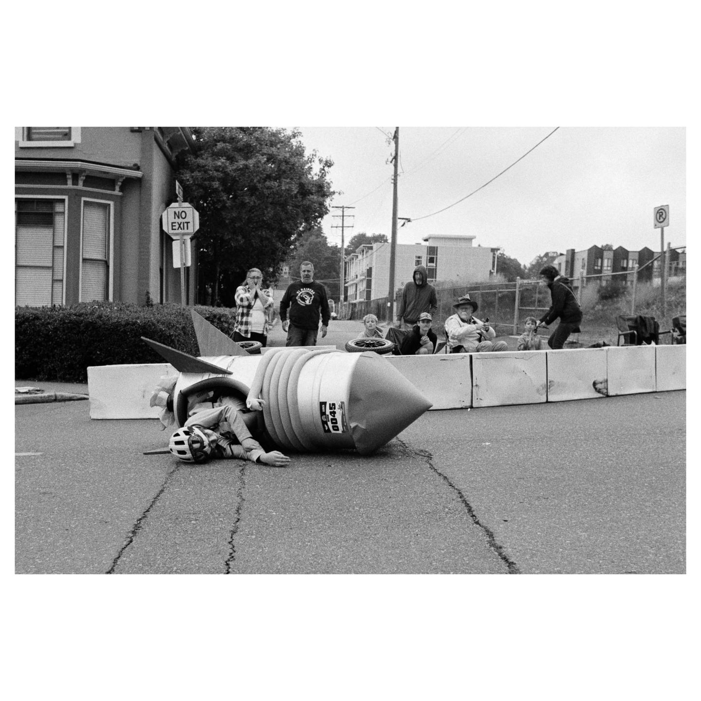 A person in a rocket-shaped soapbox car lies on the street after crashing, while spectators watch from behind a barrier.