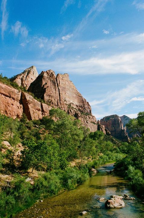 A river flows through a valley surrounded by large rock formations and green trees under a blue sky with clouds.
