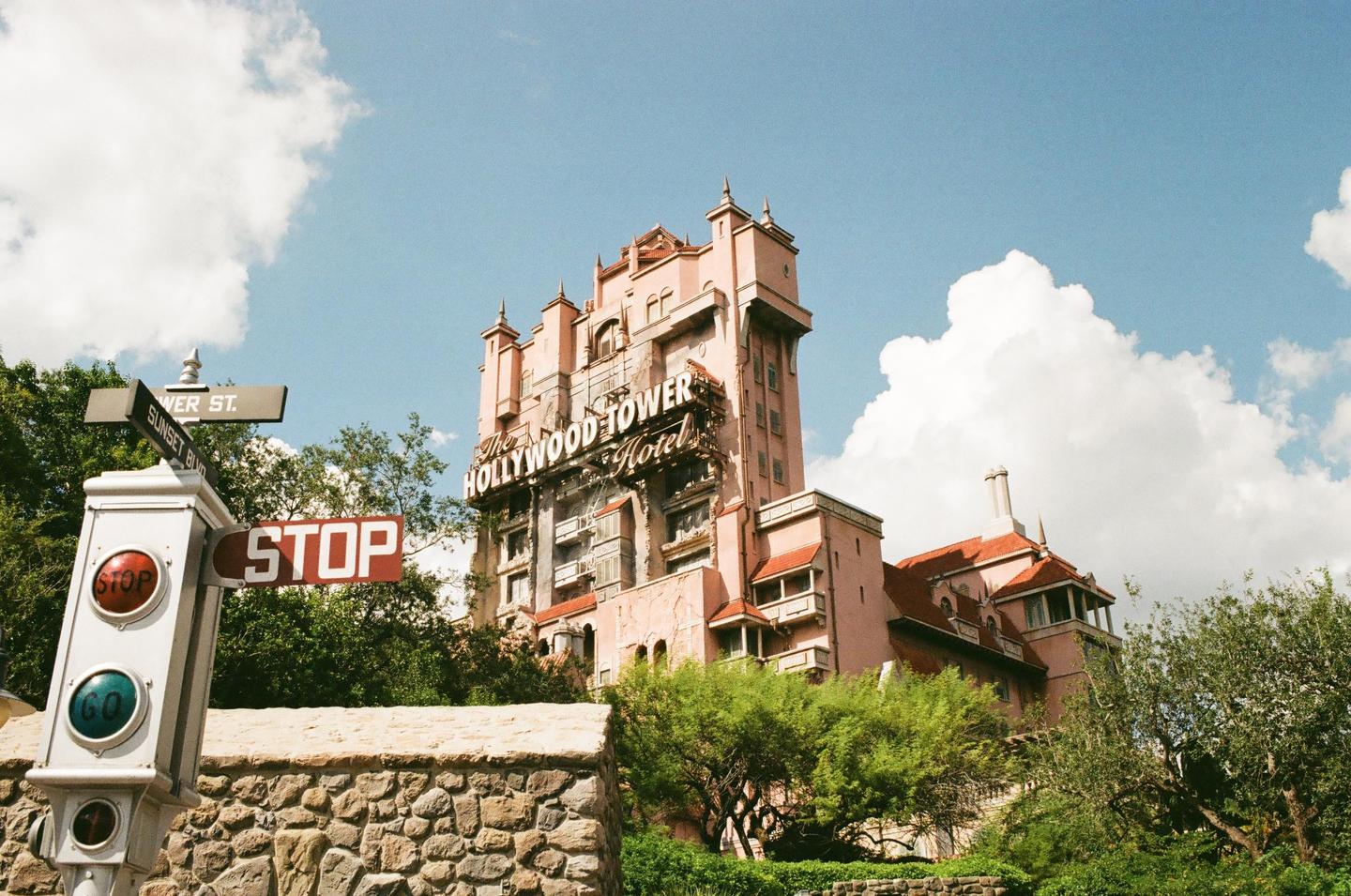A pink hotel with a red roof stands against a blue sky with white clouds, with a stop sign and street signs in the foreground.