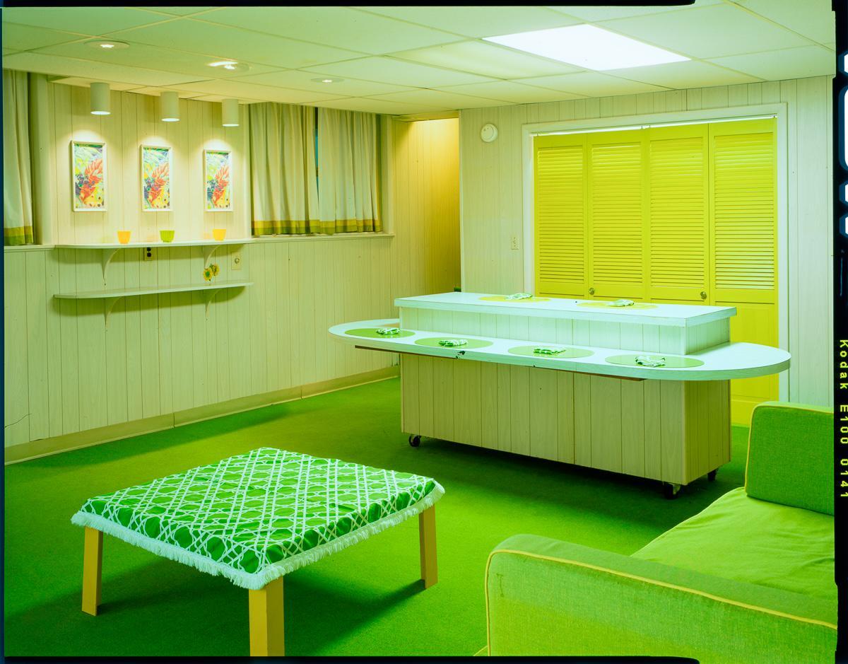 A room with green carpet and furniture features a long counter with place settings and a table with a patterned tablecloth.