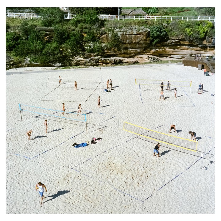 People play volleyball on a sandy beach with cliffs and green foliage in the background.