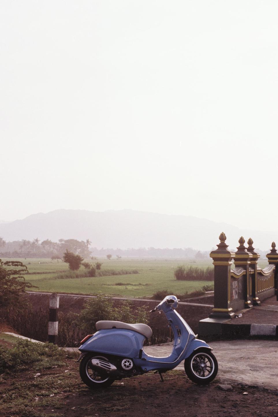 A blue scooter is parked on a dirt road next to a bridge with a rice field and mountains in the background.