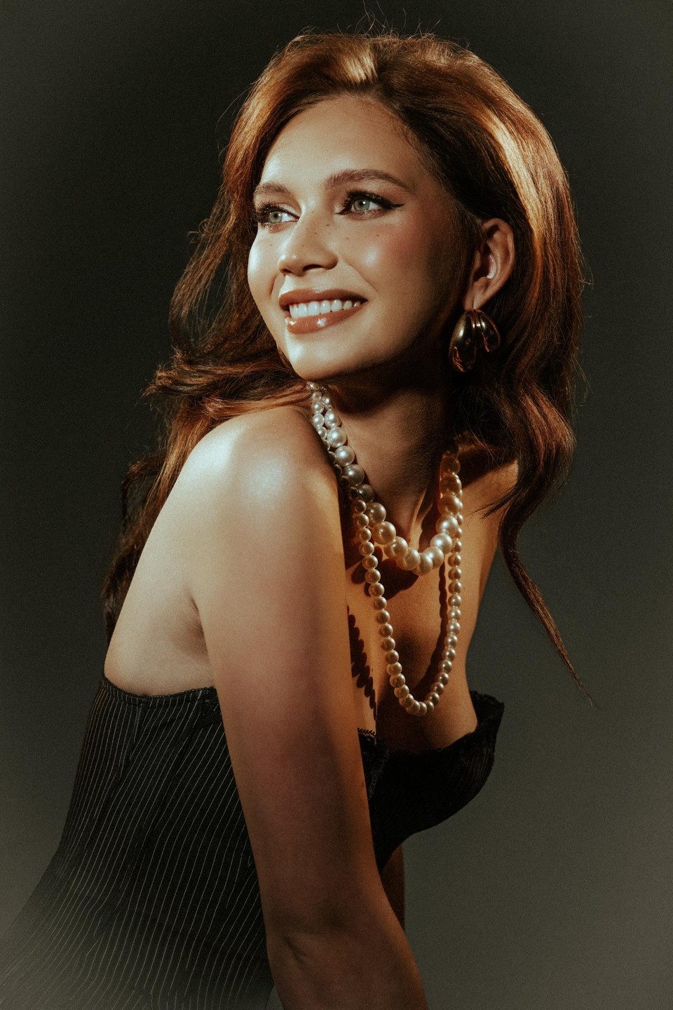 A woman with long hair and pearl necklaces smiles while looking to the side.
