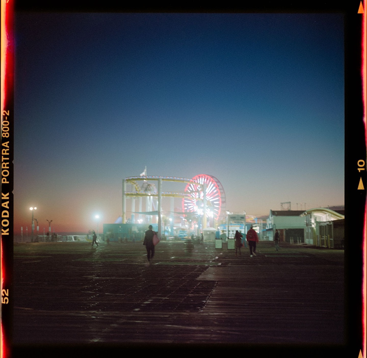 People walk on a pier towards a brightly lit Ferris wheel and roller coaster at dusk.