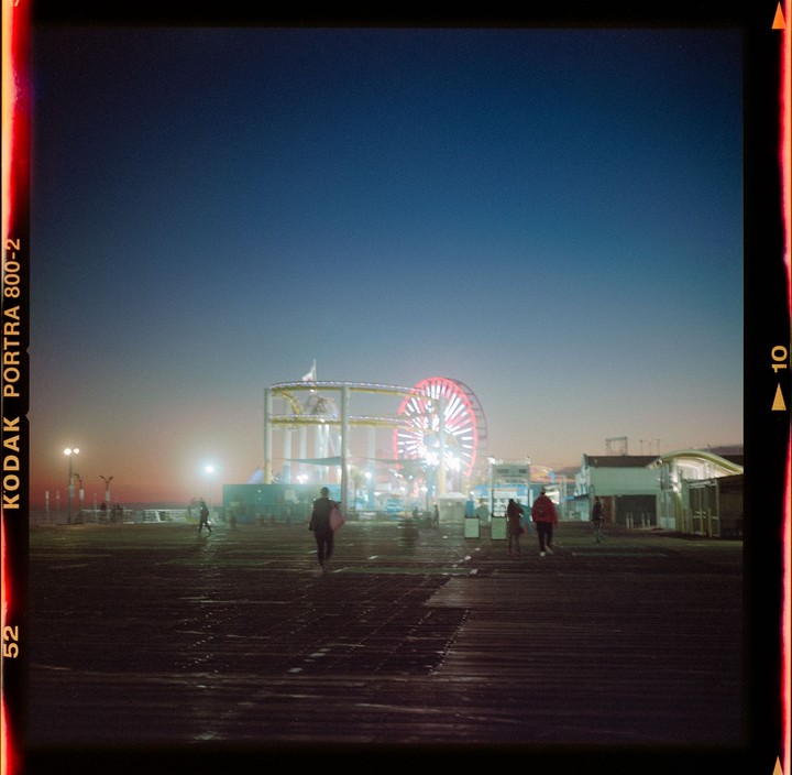 People walk on a pier towards a brightly lit Ferris wheel and roller coaster at dusk.