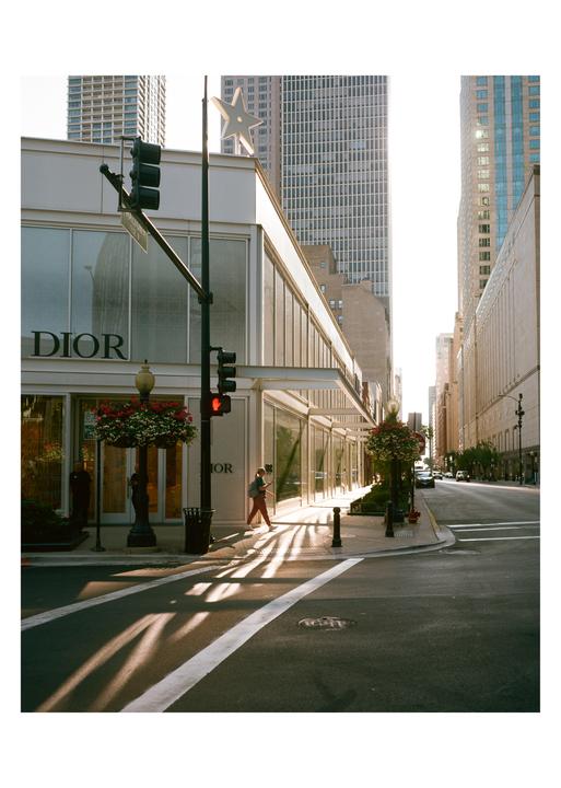 A woman walks on a sunlit sidewalk past a Dior store in a city.