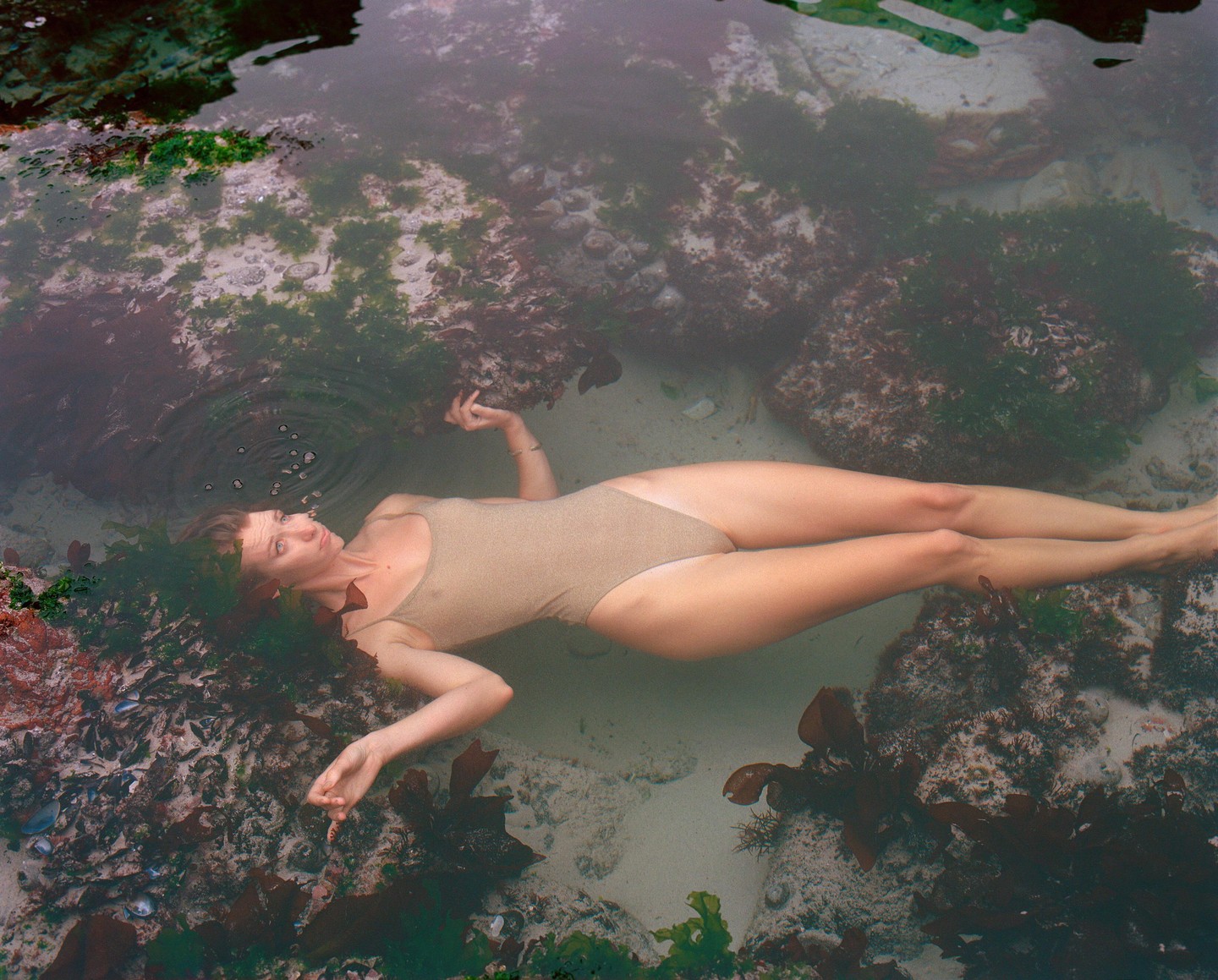 A woman floats on her back in a rock pool surrounded by seaweed.