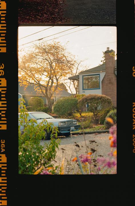 A light blue car is parked on a residential street with houses and autumn trees in the background.