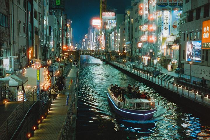 A boat full of people cruises down a canal at night, surrounded by brightly lit buildings and walkways.