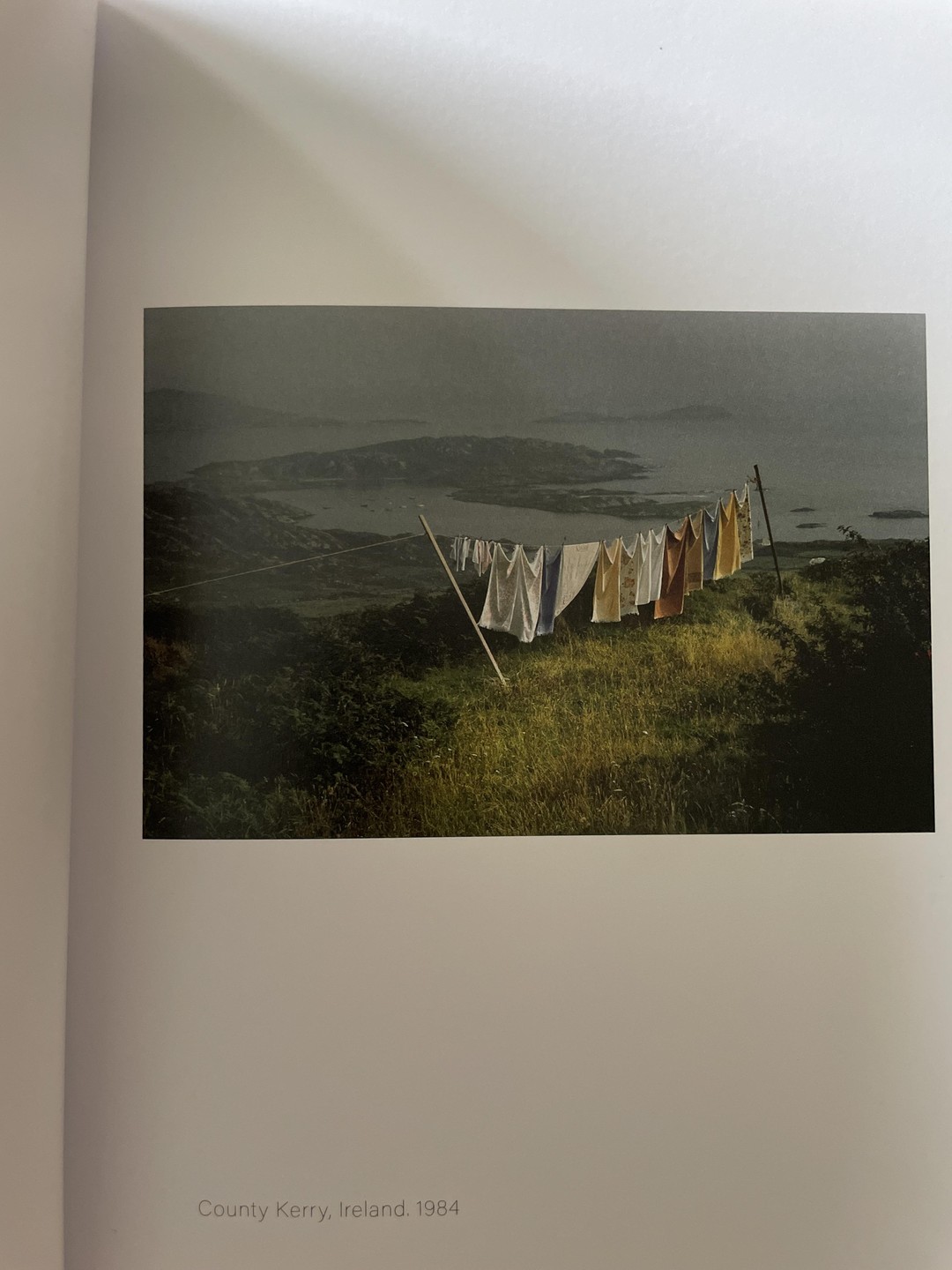 Laundry hangs on a clothesline on a grassy hill overlooking a bay with islands under a hazy sky.