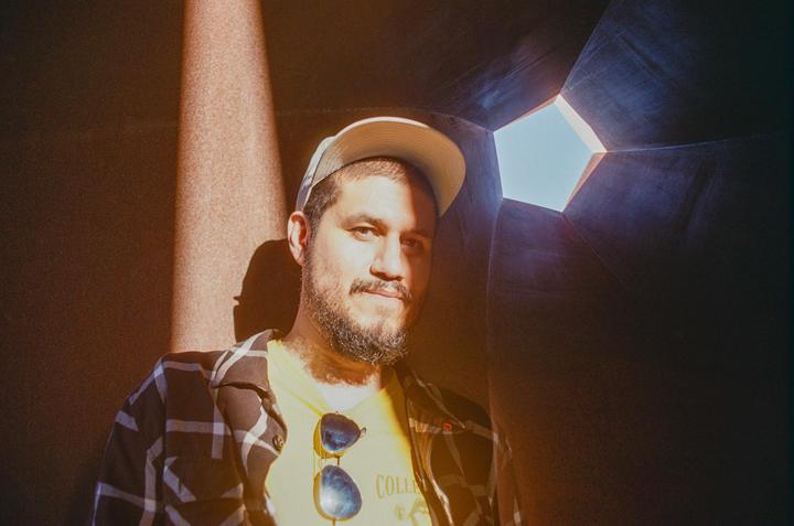 A man wearing a cap and sunglasses on his shirt is illuminated by a shaft of light in a dark, angular space.