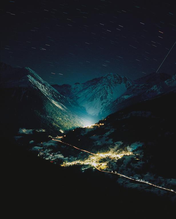 A small village with illuminated buildings and roads sits nestled in a snowy valley surrounded by mountains under a night sky with star trails.