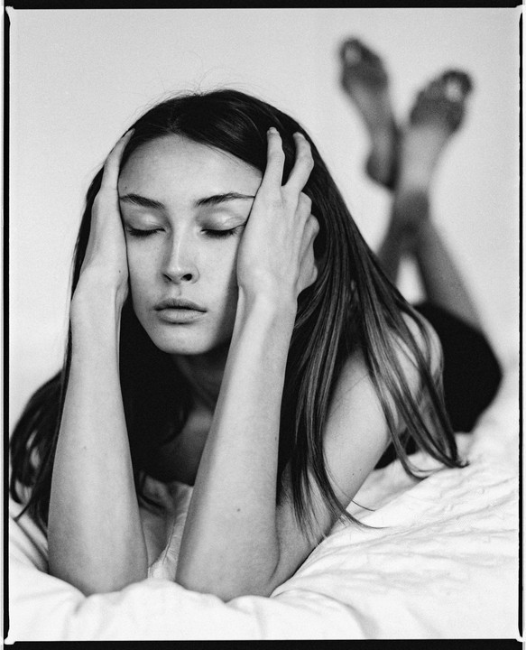 A woman lies on a bed with her eyes closed and hands on her head, with her feet crossed in the background.