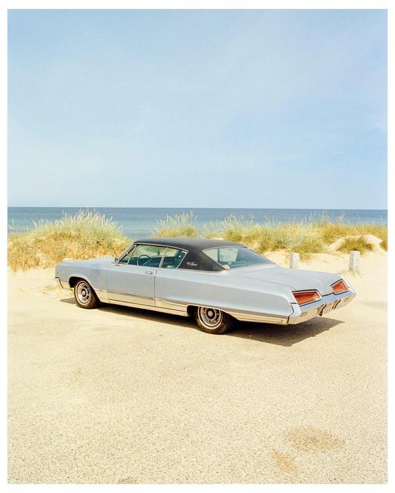 A light blue vintage car with a black roof is parked on a sandy road next to a beach with tall grass and the ocean.