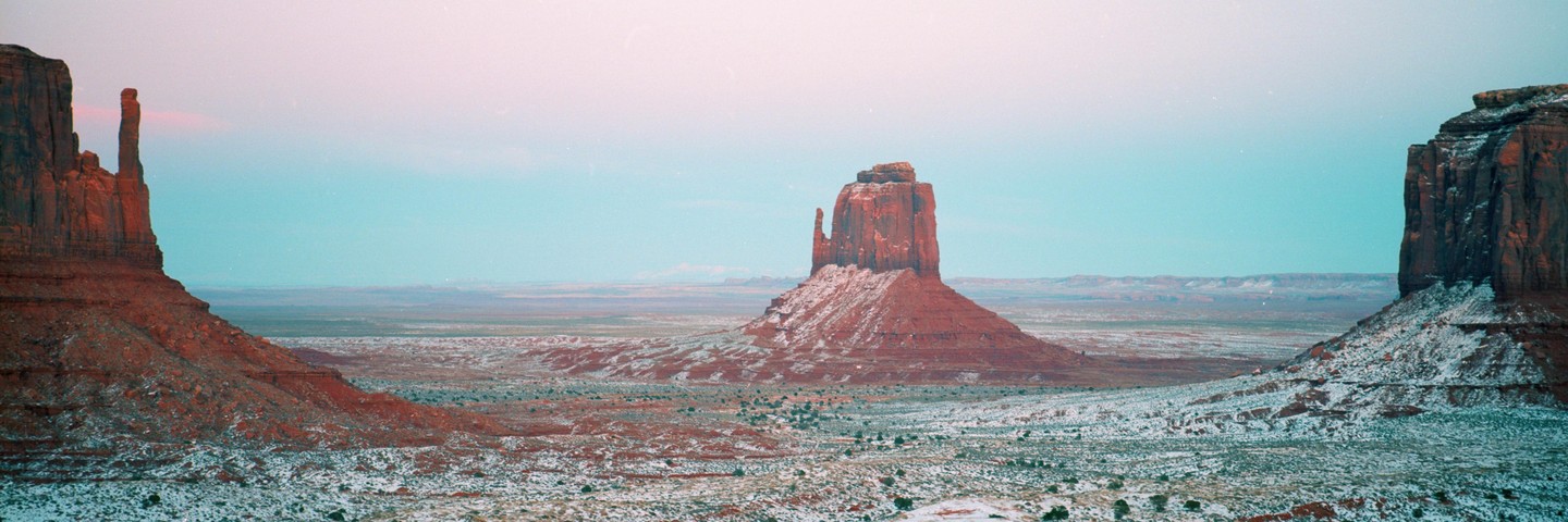 A panoramic landscape shows a snow-dusted desert valley with large red rock formations under a blue sky.