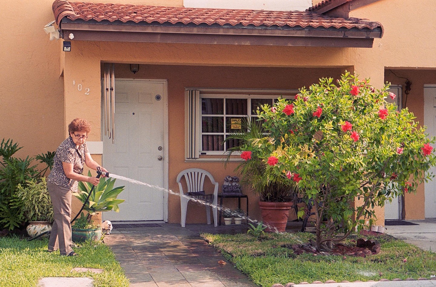A woman waters plants in front of a house with a small dog peeking out from behind a plant.