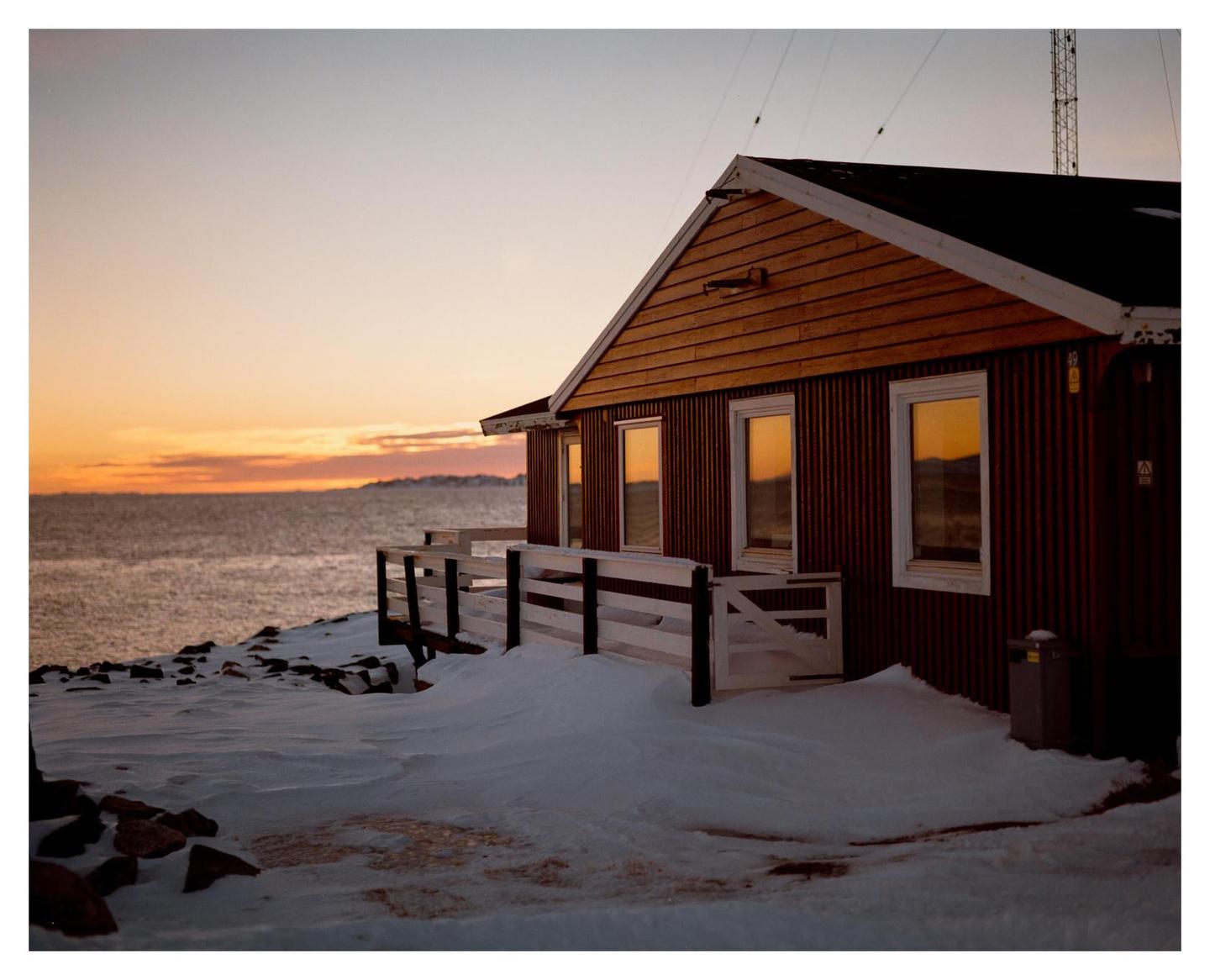 A wooden house with white trim sits on a snow-covered shore next to a body of water at sunset.
