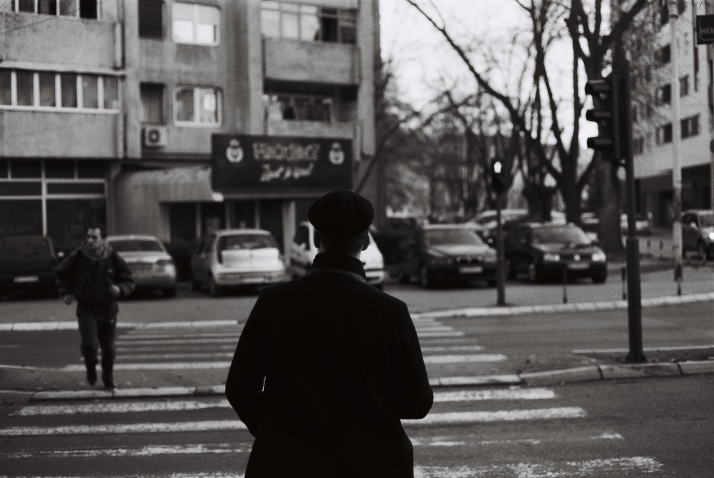 A man in a flat cap stands at a crosswalk with his back to the viewer, while another man walks across the street in the background.