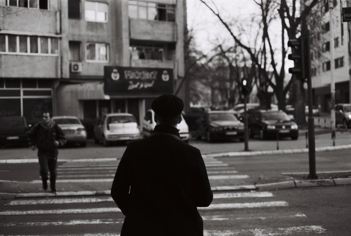 A man in a flat cap stands at a crosswalk with his back to the viewer, while another man walks across the street in the background.