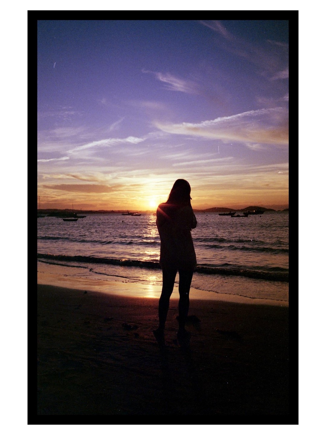 A woman stands on a beach looking out at the ocean during sunset.