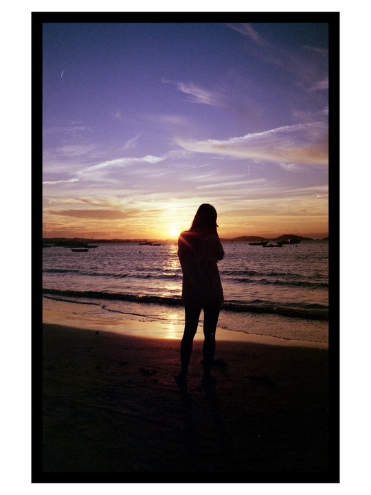 A woman stands on a beach looking out at the ocean during sunset.