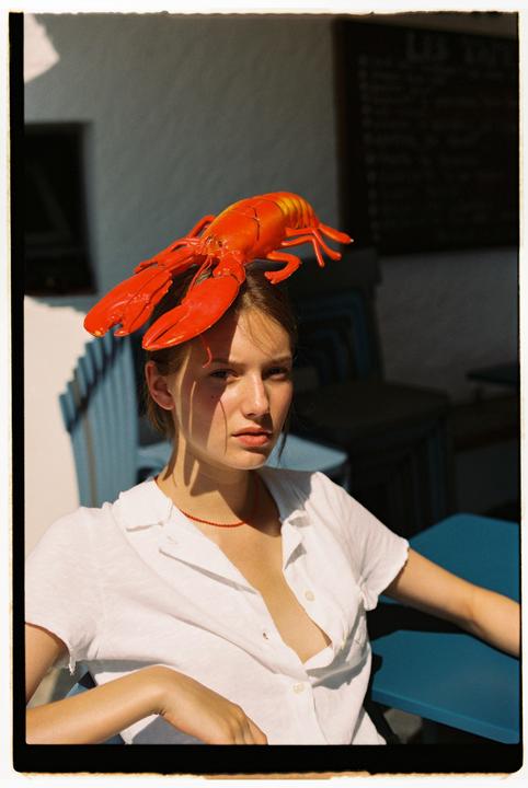 A woman with a toy lobster on her head sits at a blue table in dappled sunlight.