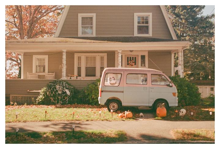 A two-tone van is parked in front of a house decorated for Halloween with pumpkins and skeletons.