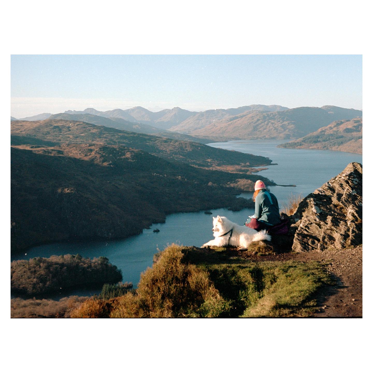 A woman and a white dog sit on a grassy cliff overlooking a lake and mountains.